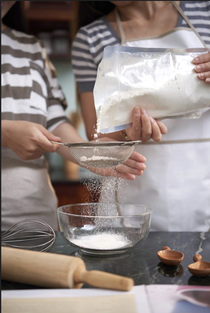 Two women in a kitchen cooking. One women is pouring flour in a bowl while the other one is helping by filtering the flour